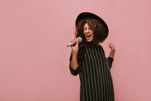 emotional-woman-with-curly-brunette-hairstyle-stylish-hat-striped-black-dress-holding-microphone-singing-pink-wall-min